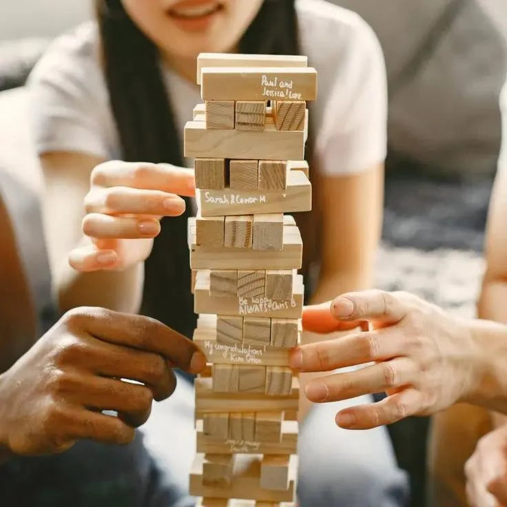 A close-up of a wooden block stacking game being played by multiple people, with hands reaching for the tower and a blurred indoor background.