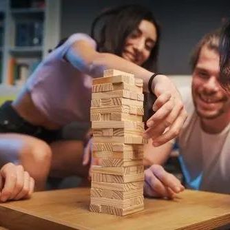 Two people playing with a wooden block stacking game on a wooden table in a dimly lit room.